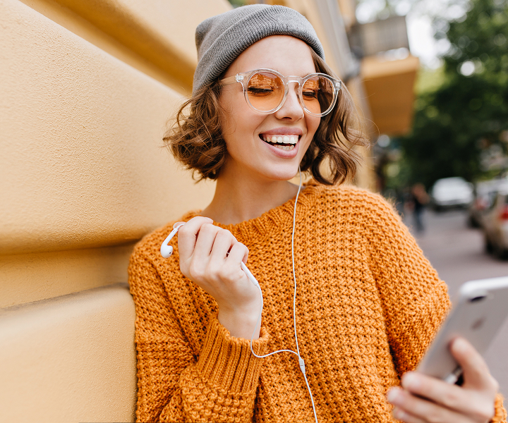 Smiling person with phone and earbud outdoors.